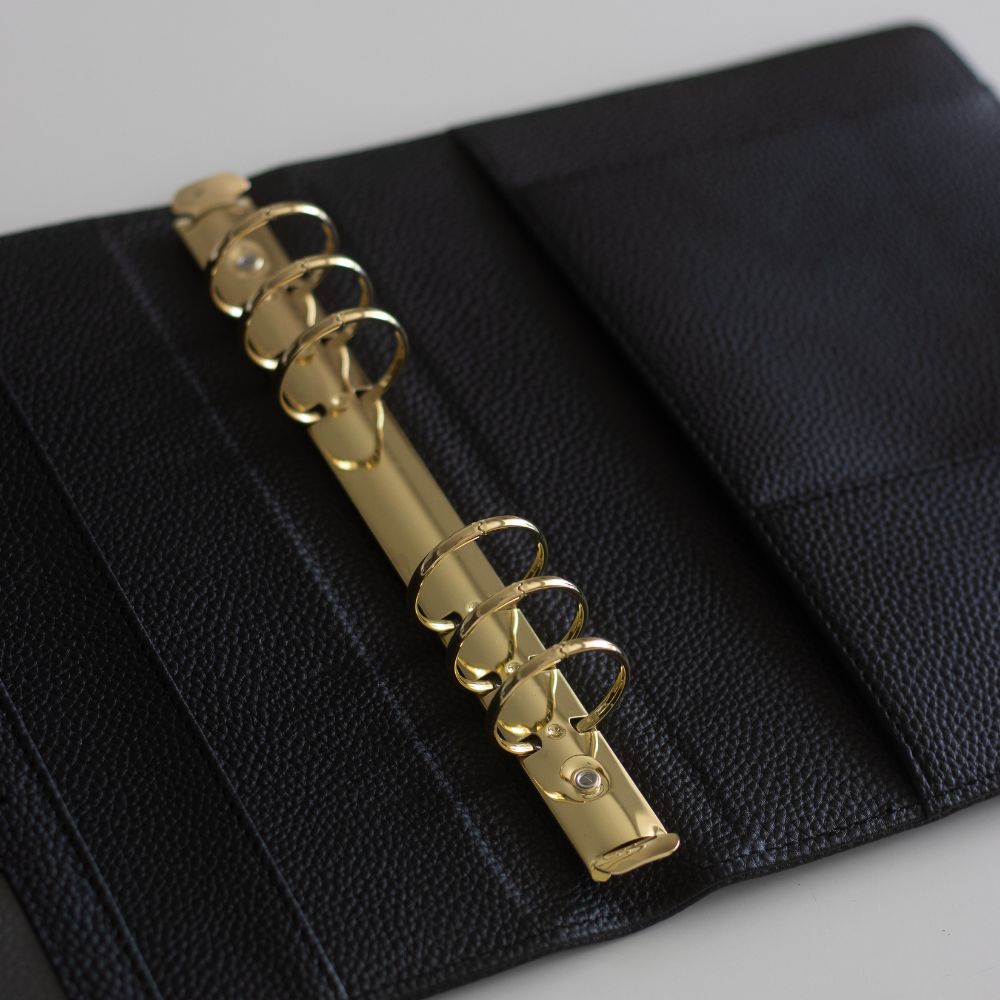Black textured vegan leather ringbinder cover with six gold rings on a light grey background