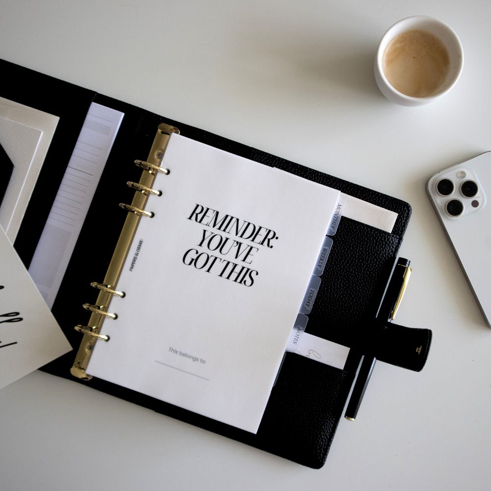 Black A5 planner with a motivational quote on a white surface with a cup of coffee and phone.