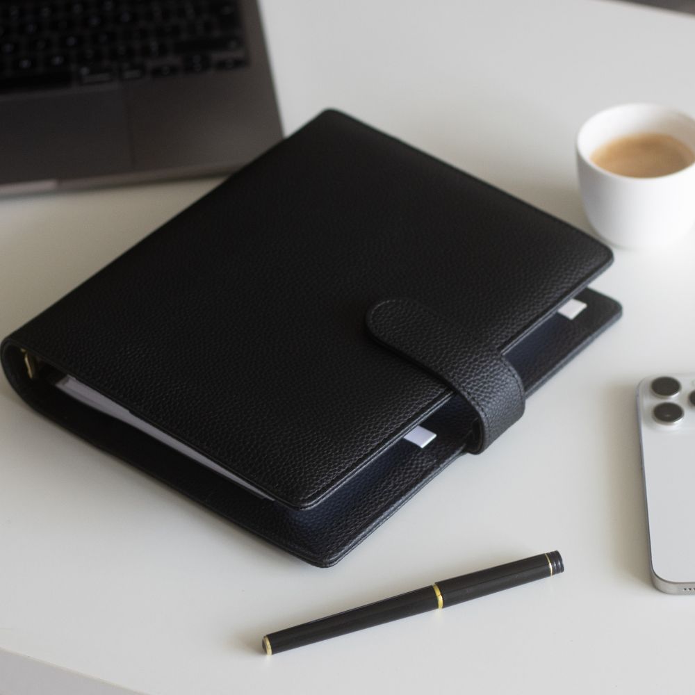 Black A5 vegan leather planner on a desk with a pen, smartphone, and coffee cup.