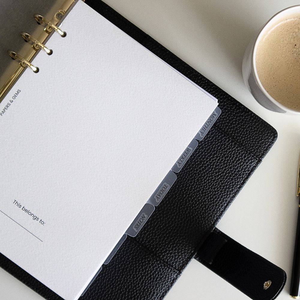 Black leather ringbinder planner with white pages on a light surface, next to a cup of coffee