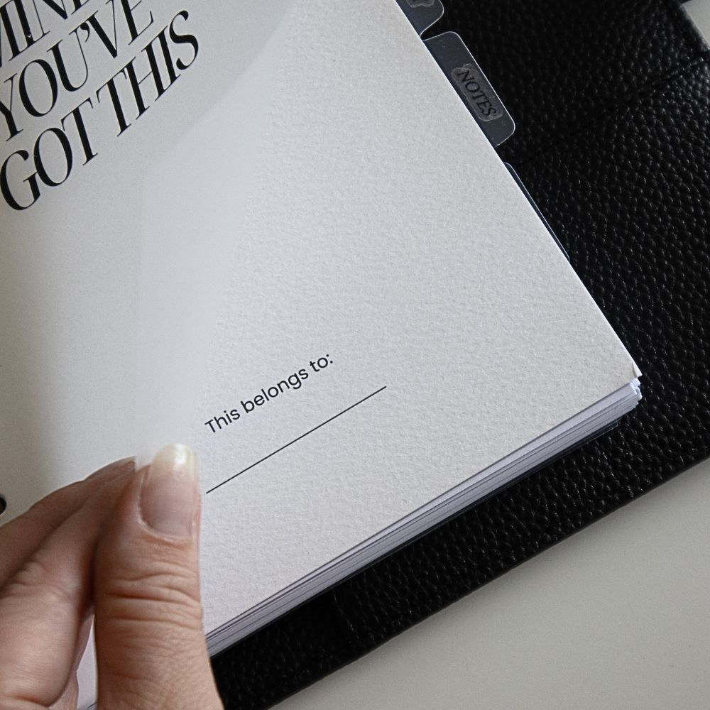 Black faux leather ringbinder planner with a dashboard page titled 'This belongs to' held by a hand.