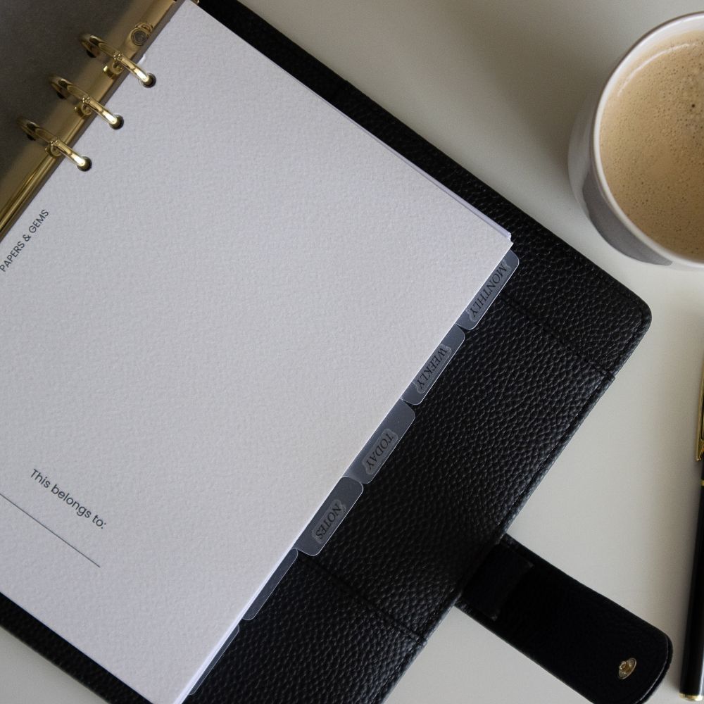 Black textured vegan leather ringbinder cover with white pages on a light surface, next to a cup of coffee.