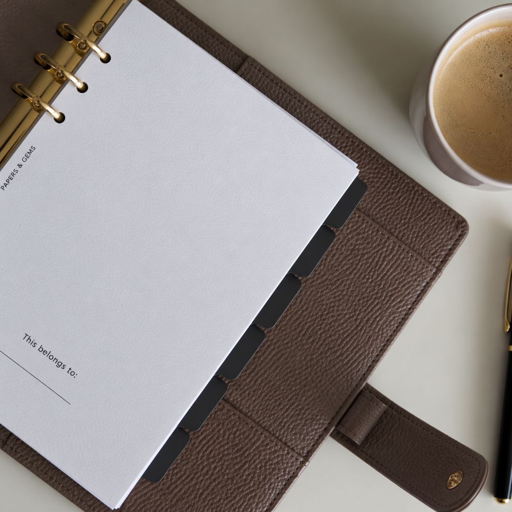 A brown 6-ring pebbled leather Agenda planner cover holding a planner, with gold rings, shown on a light neutral desk with a cup of coffee and a pen nearby.