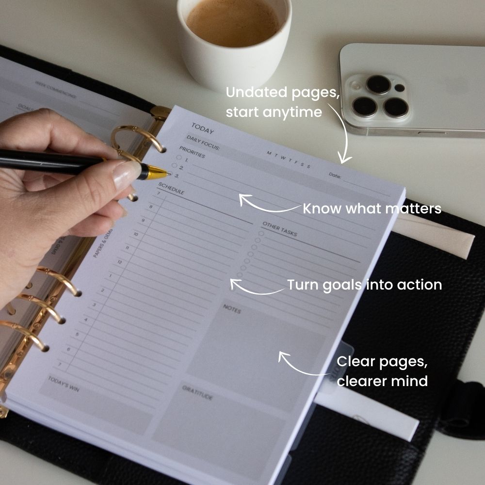 An open black leather-bound planner with gold rings, showing white undated daily planning pages. A hand holds a black pen over the page.
